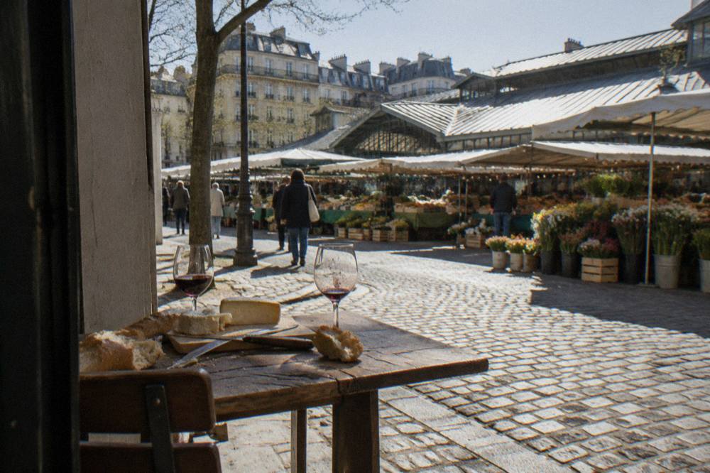 Cheese, bread and glasses of wine arranged on a wooden table beside an open market street