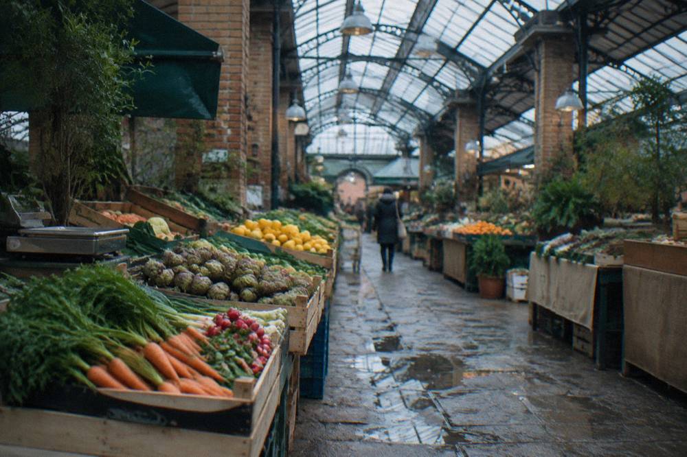Narrow covered market hall with produce stalls and shoppers under iron and glass roofing