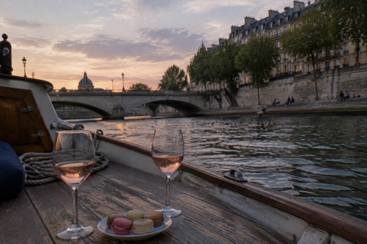 Small private boat on the Seine at dusk passing stone bridges, glasses of rosé and macarons on a wooden deck table