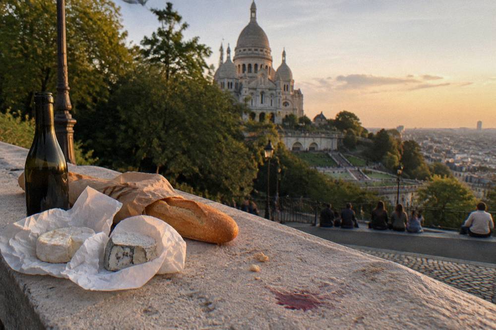 A baguette, cheeses and a bottle of wine arranged on a park bench for a picnic