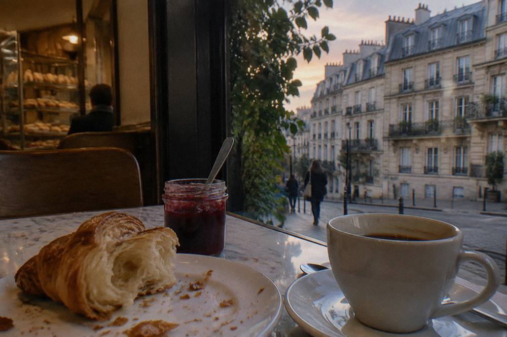 A torn croissant on a small plate beside a cup of black coffee and a jar of jam