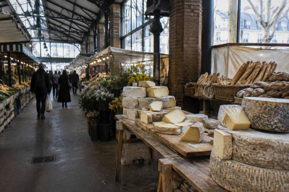 Cheese counter with stacked wheels and wedges beside a bread stall in a covered market